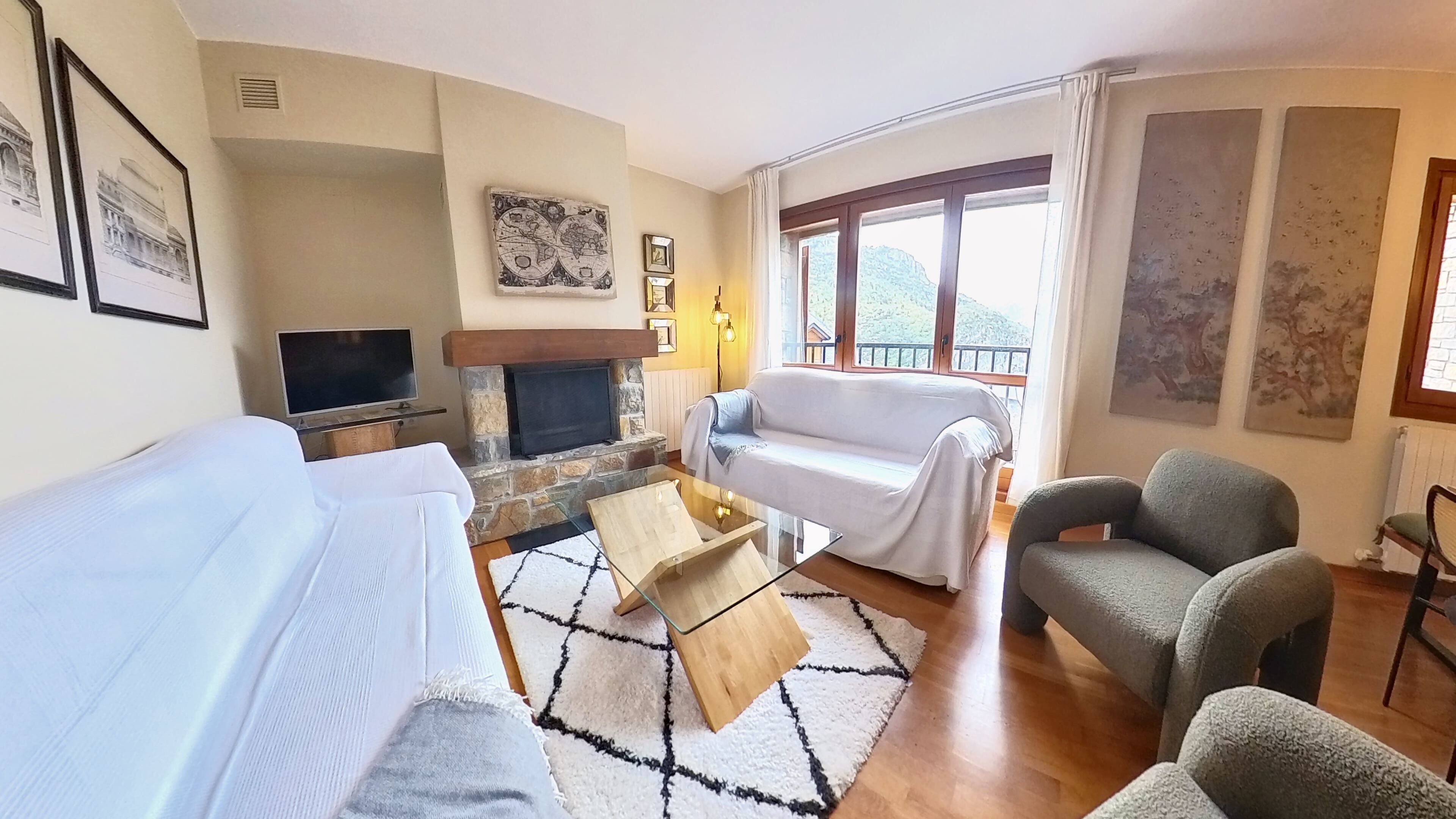 Ein Wohnzimmer mit einer Couch, einem Sessel und einem Kamin. Ein Fernseher und ein Fenster mit Blick ins Freie sind ebenfalls vorhanden.