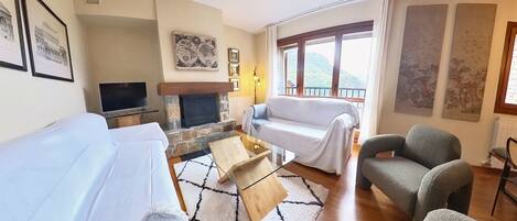 Ein Wohnzimmer mit einer Couch, einem Sessel und einem Kamin. Ein Fernseher und ein Fenster mit Blick ins Freie sind ebenfalls vorhanden.