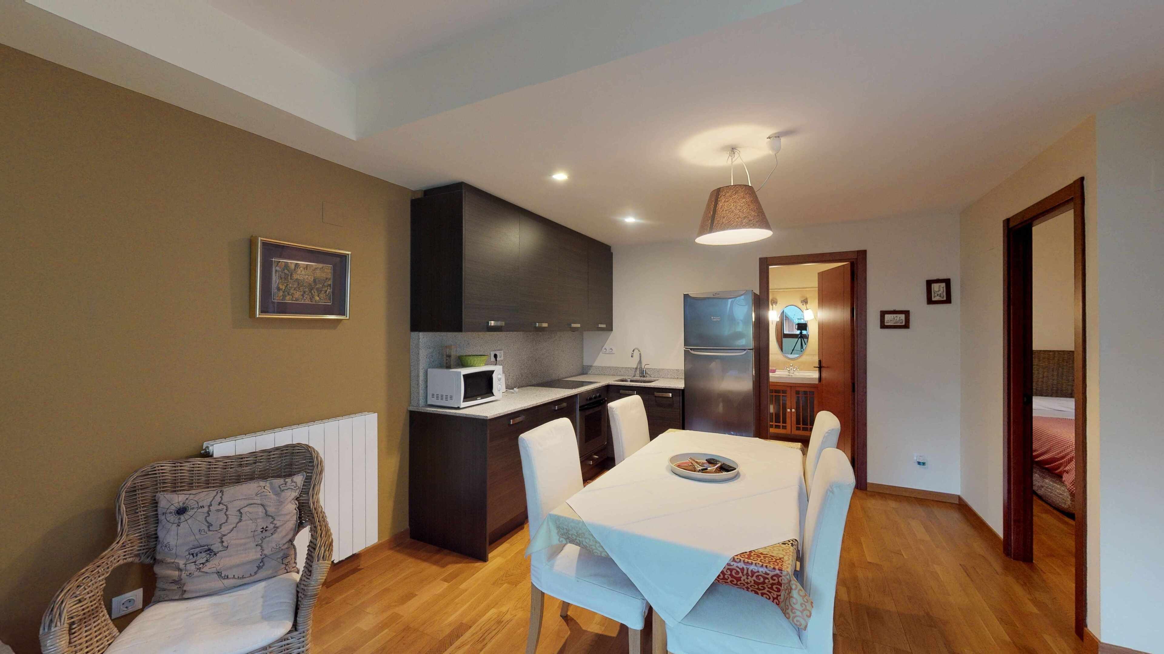 A compact kitchen with dark cabinets, a microwave, and a refrigerator. There is a dining table with chairs and a wicker chair with a map cushion.