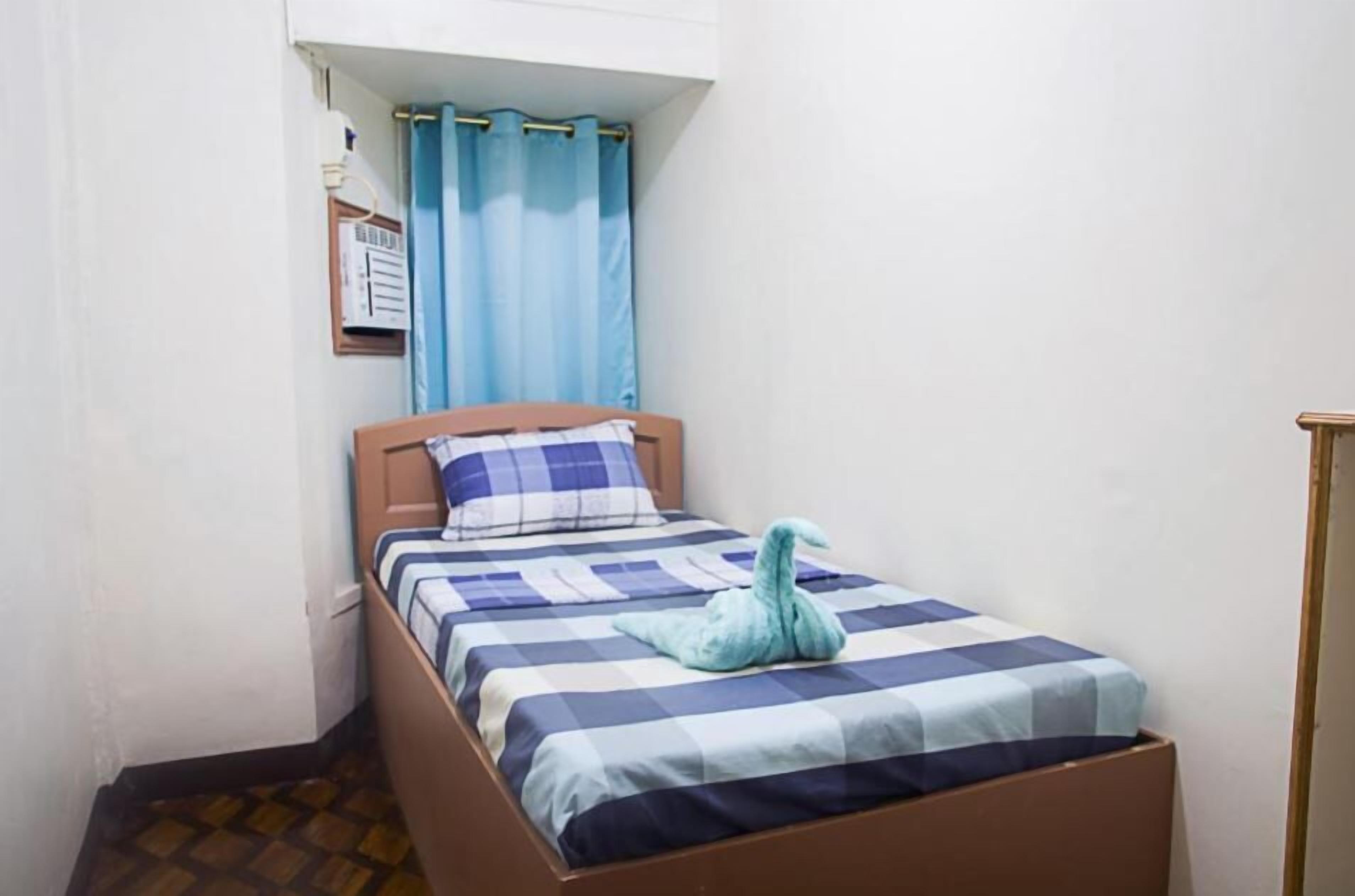 Une petite chambre avec un lit simple, une couvre-lit à carreaux, un rideau bleu et un luminaire fixé au mur.