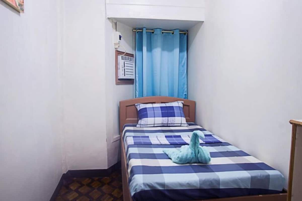 Una habitación pequeña con cama individual, colcha de cuadros, cortina azul y aire acondicionado instalado en la pared.