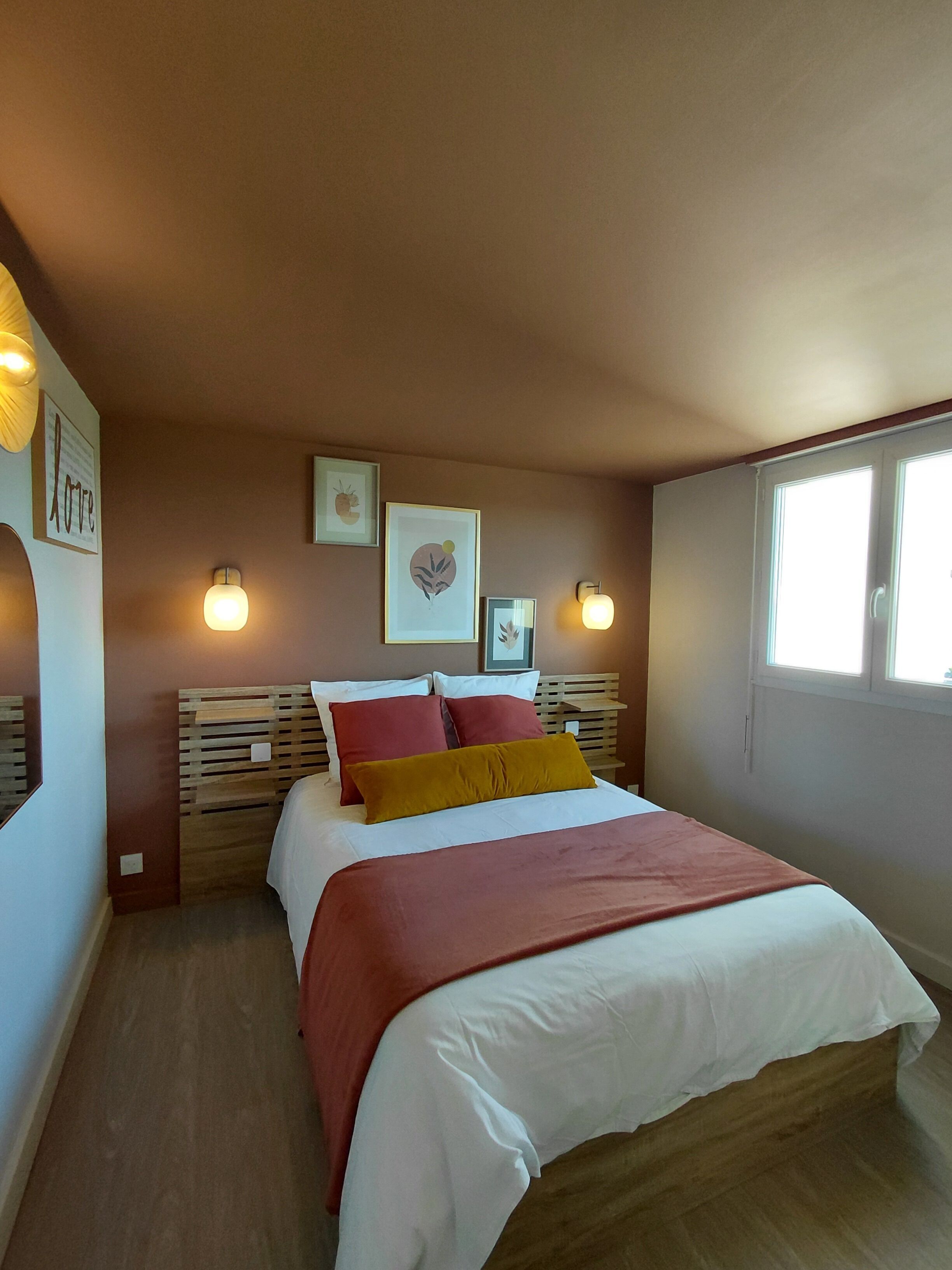 A bedroom with a bed, wooden headboard, wall-mounted lights, framed artwork, and a window.