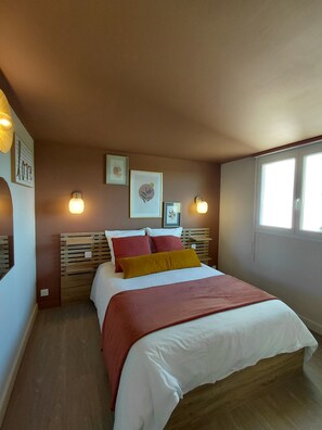 Une chambre à coucher avec un lit, une tête de lit en bois, des lampes fixées au mur, des œuvres d’art encadrées et une fenêtre.