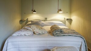 Una cama bien hecha con un edredón estampado con motivos florales, almohadas y una manta a juego. Hay dos lámparas de noche y una mesita de noche con un libro y una botella.