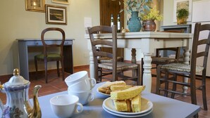 Ein Esstisch mit Teekanne, Tassen und Sandwiches. Um den Tisch herum stehen Stühle, und im Hintergrund ist eine Küche erkennbar.