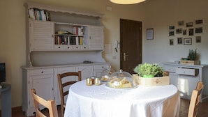 Un coin repas avec une table ronde recouverte d’une nappe blanche, entourée de chaises en bois. On y trouve une bibliothèque avec des livres et une petite plante posée sur la table.