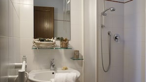 Une salle de bain avec un lavabo blanc, une étagère en verre où sont rangés les articles de toilette, un miroir et un espace douche.