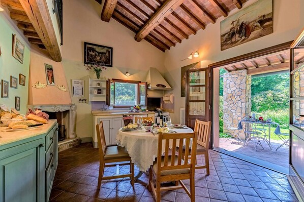 A cozy dining area with a round table set for four, wooden chairs, a kitchen area with a window, and a stone wall outside.