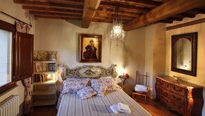 Een slaapkamer met een houten plafond, een bed met bloemenbedekking, een kroonluchter, een nachtkastje met een lamp, een dressoir en een grote spiegel.