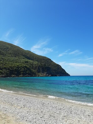 Ein Kiesstrand mit klarem türkisblauem Wasser und einem grünen Hügel im Hintergrund.
