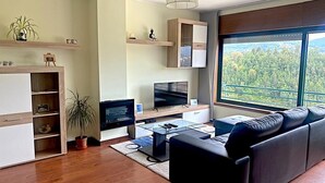 En moderne stue med et fladskærms-tv, en sofa og et spisebord. Der er træhylder og en potteplante.