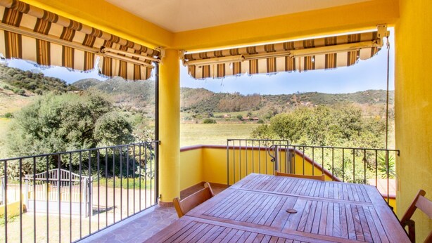 Un balcon avec une table et des chaises en bois, des murs jaunes et une vue sur un paysage vallonné.