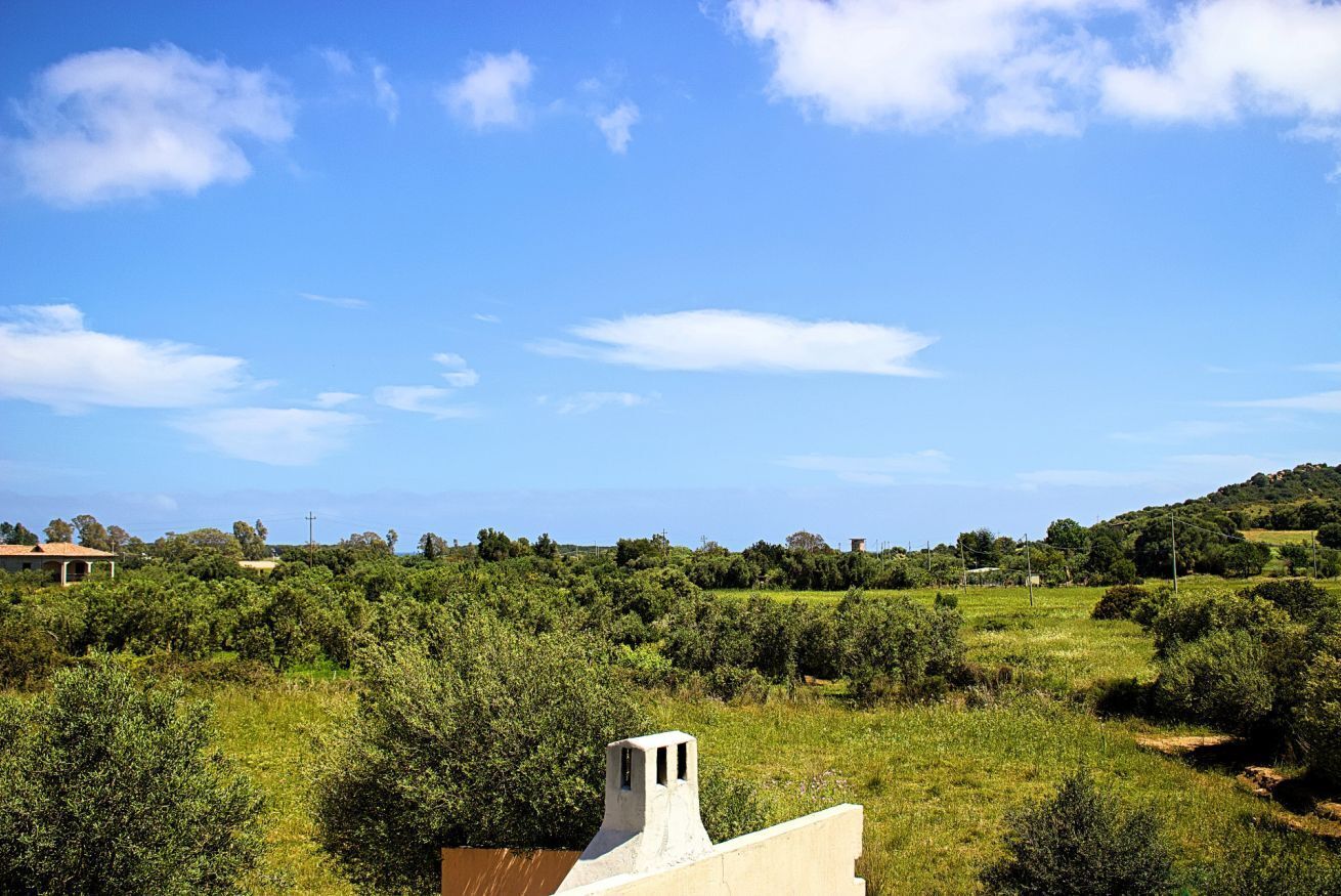 A rural landscape with a building featuring a chimney, surrounded by lush greenery and a clear blue sky.