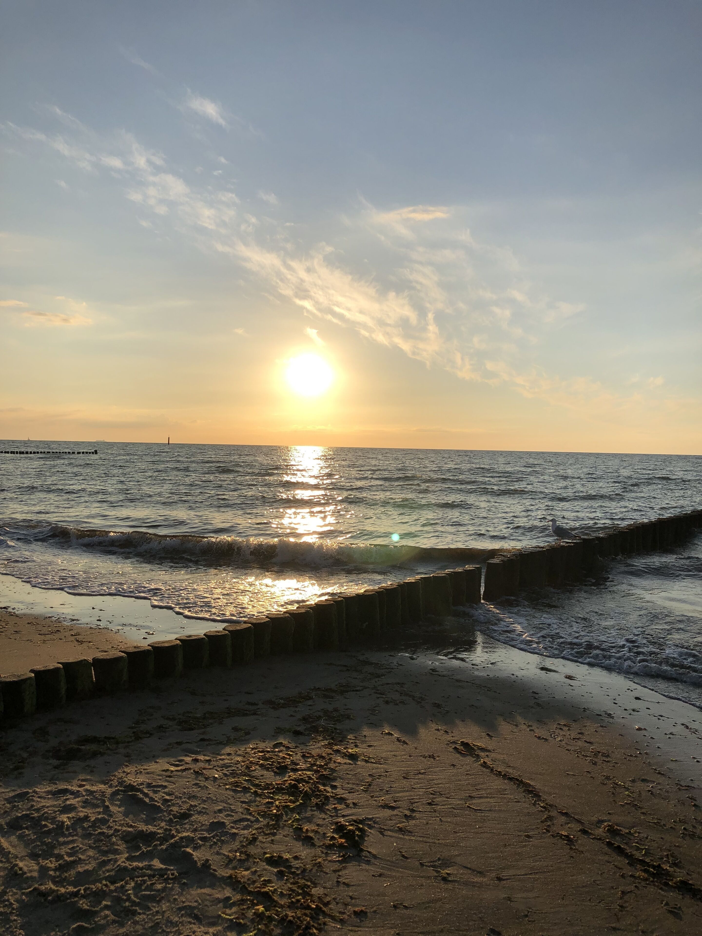 Een strand met de zon die ondergaat boven het water, golven die zachtjes op het strand komen en een houten golfbreker op de voorgrond.