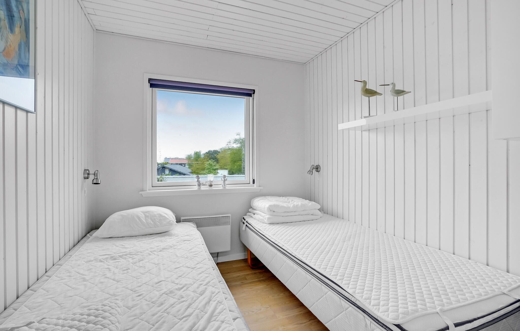 A room with two beds, a window with a view of greenery and buildings, and white walls with vertical paneling.