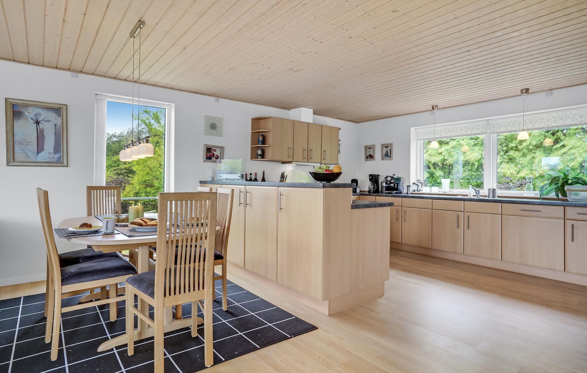 A modern kitchen with wooden cabinets, a dining area with a table and chairs, and large windows offering a view of greenery.