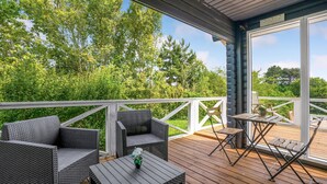 Um terraço coberto ao ar livre com móveis de vime e uma mesa, cercado por um guarda-corpo branco e vegetação exuberante.