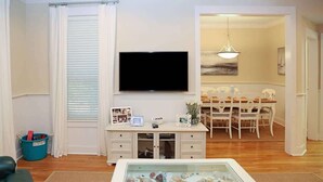Ett vardagsrum med en platt-TV, ett soffbord med glasskiva och snäckor, en matplats med ett vitt bord och stolar samt en blå papperskorg.