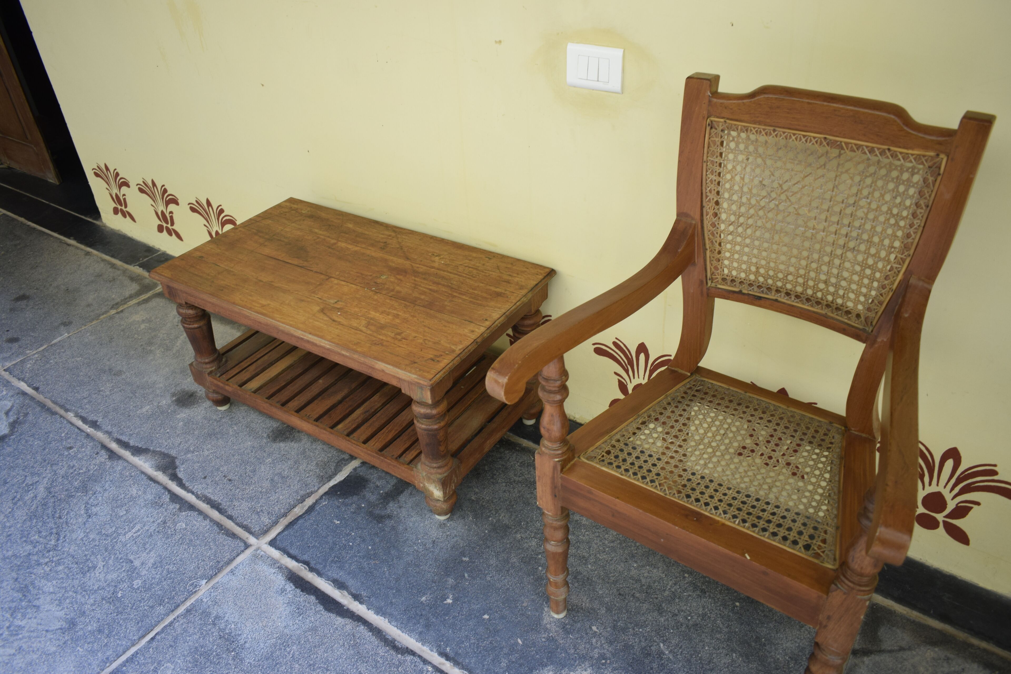 A wooden chair with a woven backrest and a matching wooden table with a slatted base, placed on a tiled floor against a wall with decorative floral patterns.