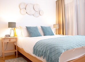 Ein ordentlich bezogenes Bett mit blauer Decke, zwei Nachttischlampen, einer Wanduhr und einem kleinen Holennachttisch.