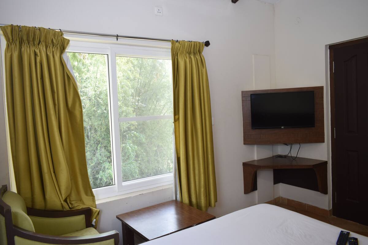 A hotel room with a bed, a chair, a TV mounted on a wooden shelf, and a window with curtains.