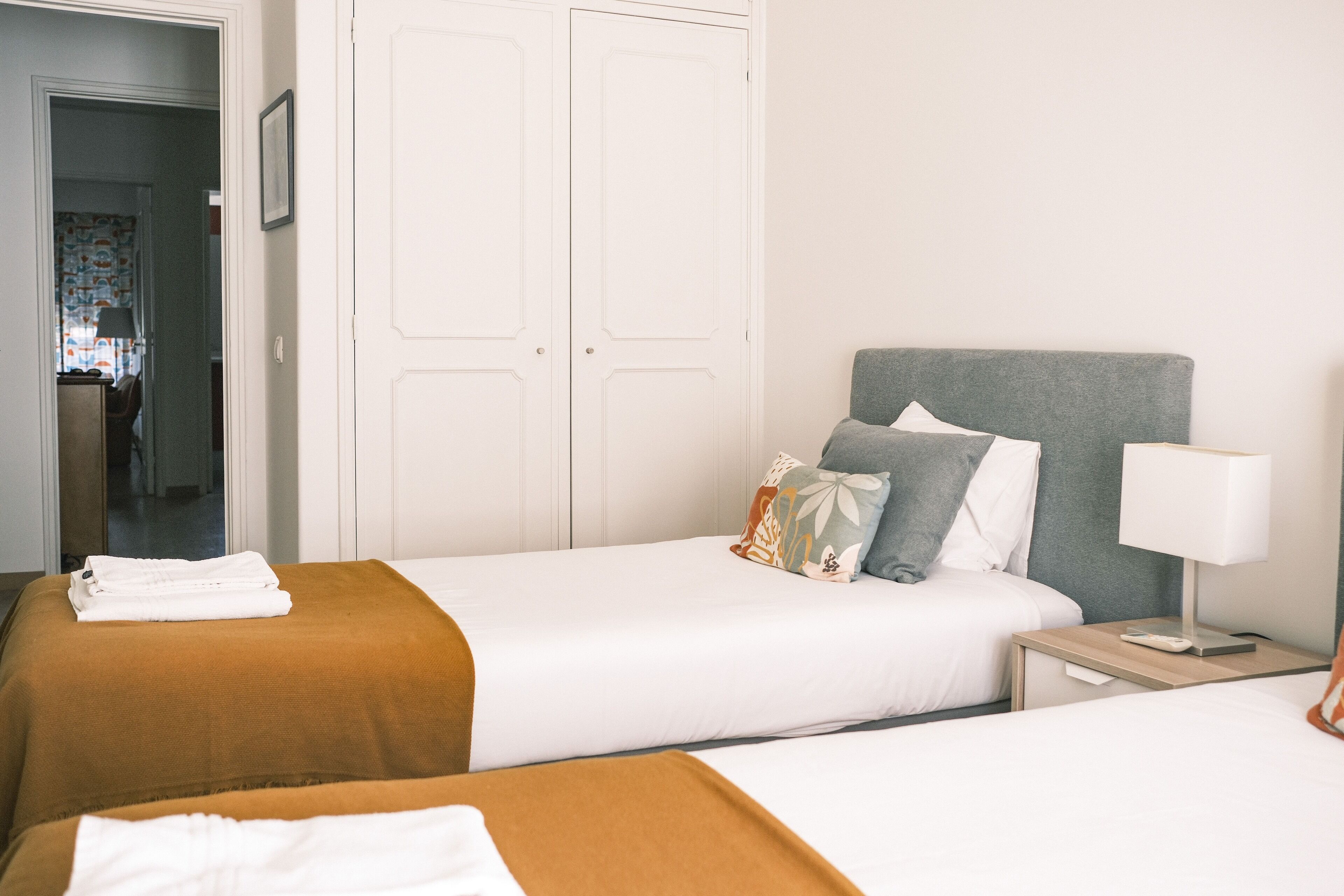 A hotel room with two beds, a grey headboard, white walls, a white door, and a small bedside table with a lamp.