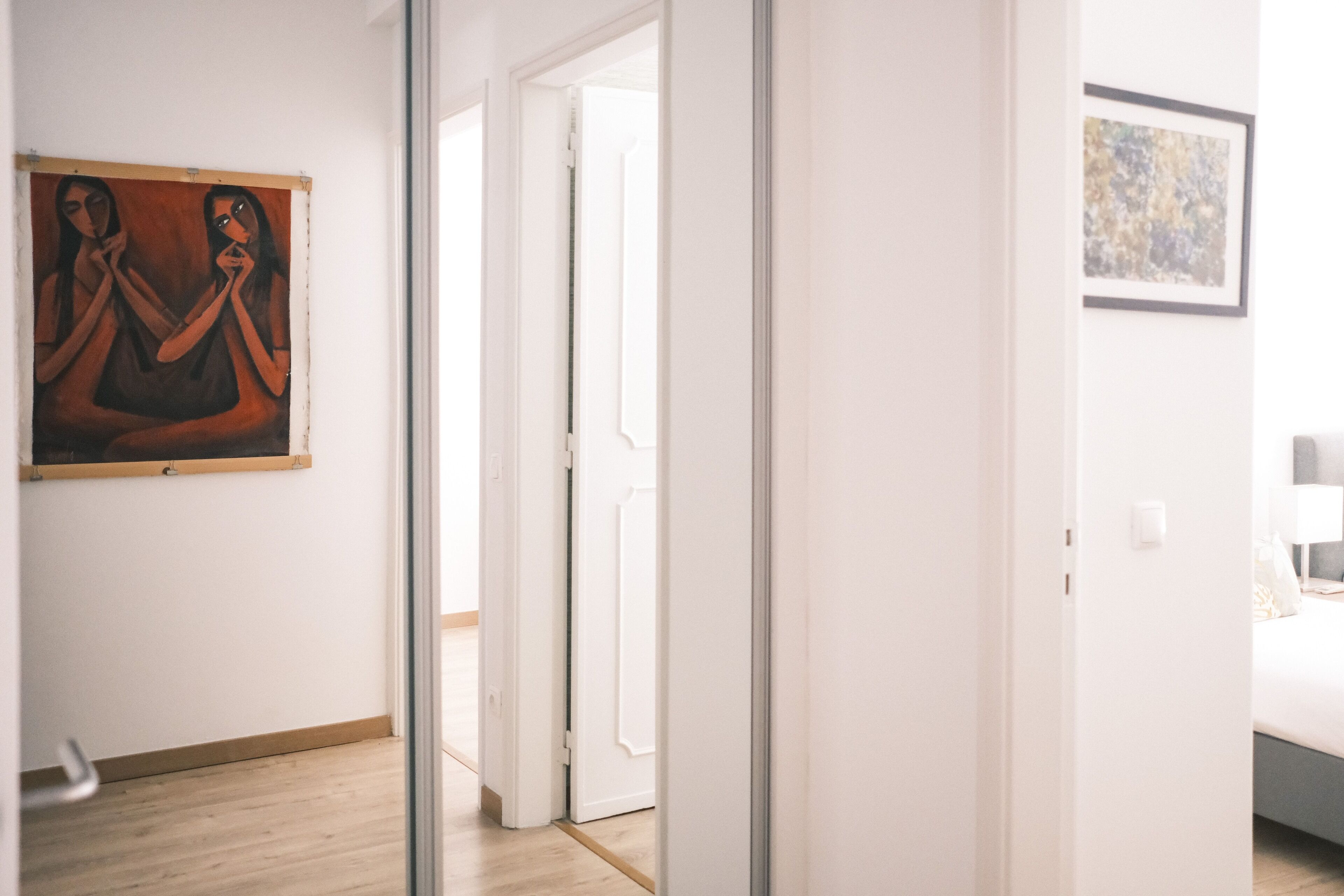 Une chambre d’hôtel avec un plancher en bois, un lit doté d’une tête de lit grise, un mur blanc orné d’un tableau abstrait encadré et une porte donnant sur une autre pièce.