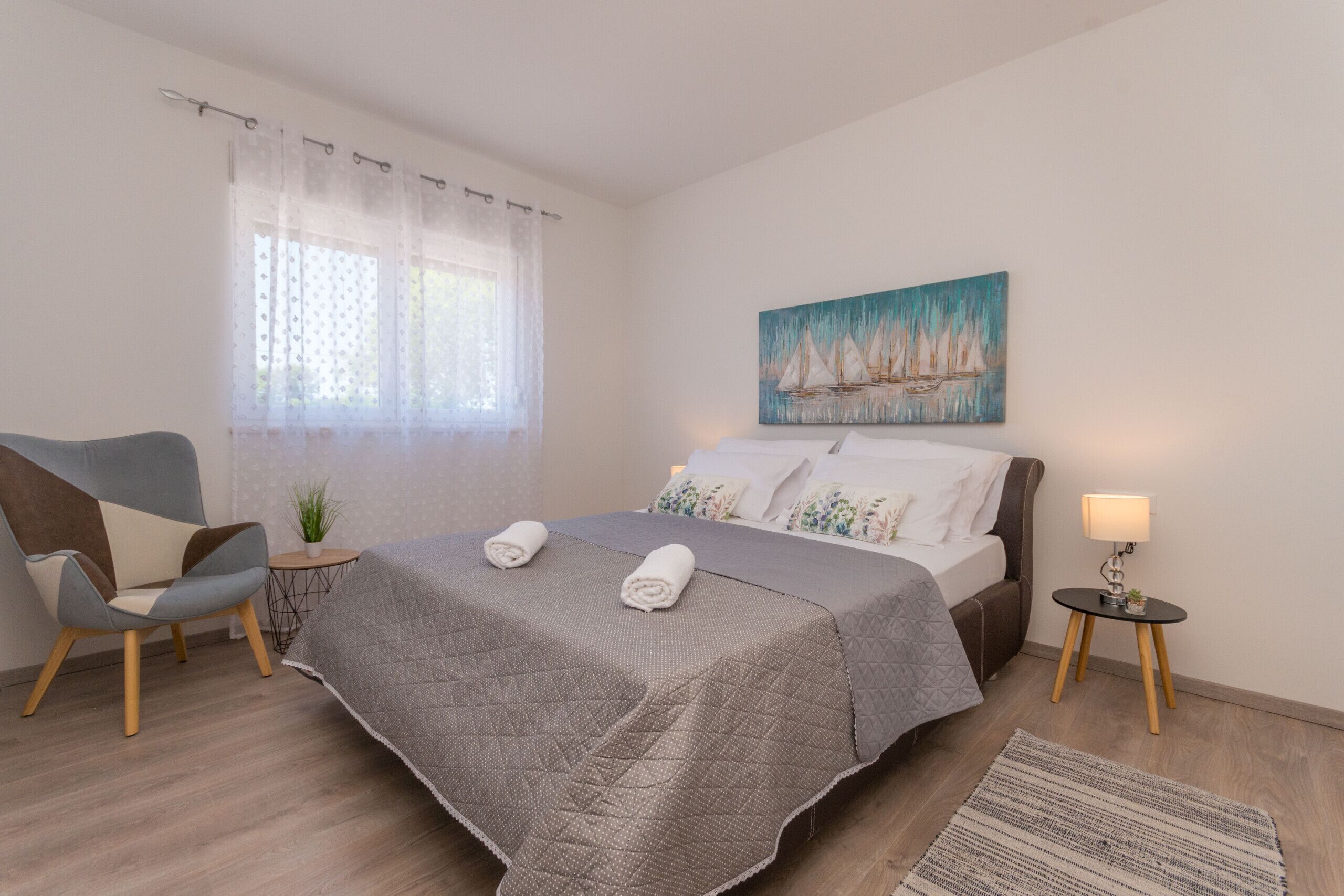 Un dormitorio ordenado con cama, una silla, una mesita y un cuadro en la pared.