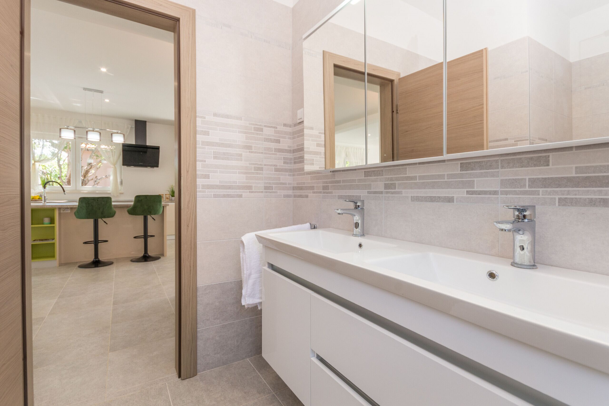 A modern bathroom with a large mirror, two sinks, and a view into the adjacent kitchen area.