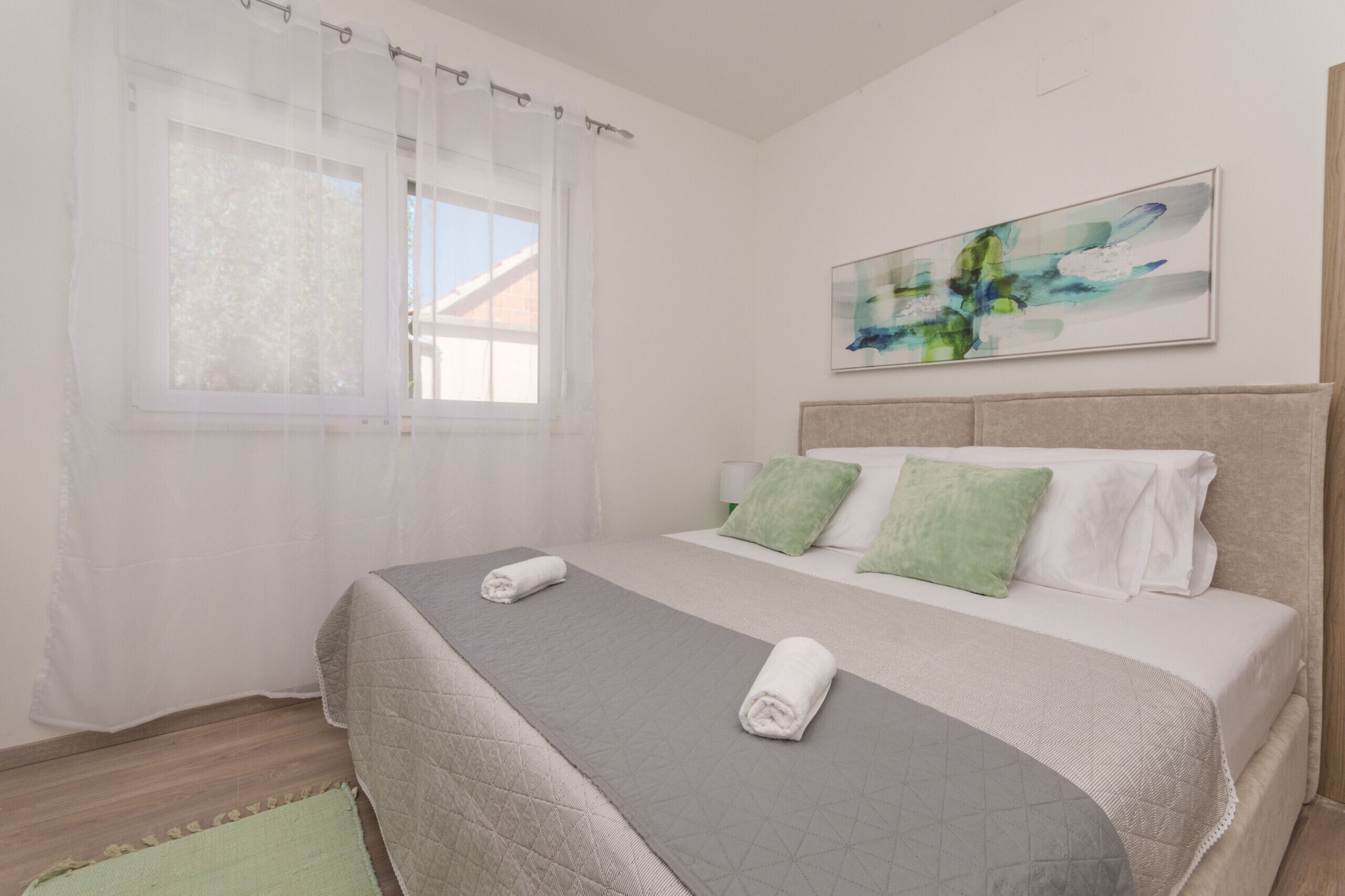 Una cama bien hecha con un edredón gris, una almohada verde y una almohada blanca. Hay un cuadro en la pared sobre la cama.