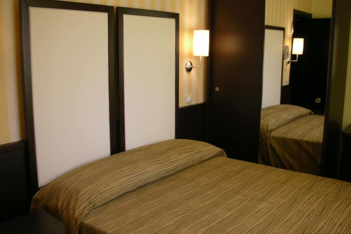 Quarto de hotel com duas camas de solteiro, cabeceira de madeira, espelho e luminária fixada na parede.