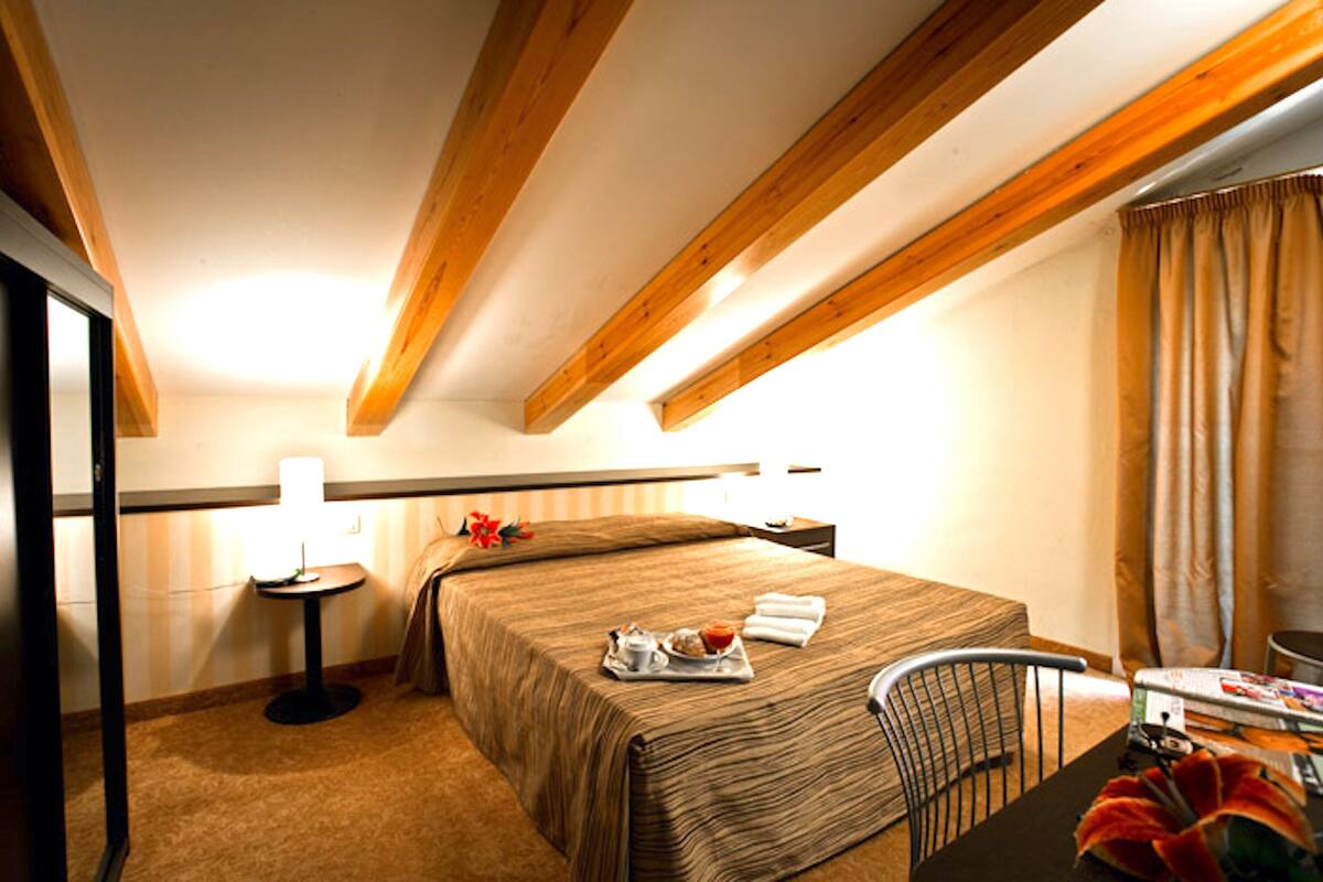 Quarto de hotel com uma cama, uma mesinha com luminária, uma cadeira e um tabuleiro com itens de café da manhã.