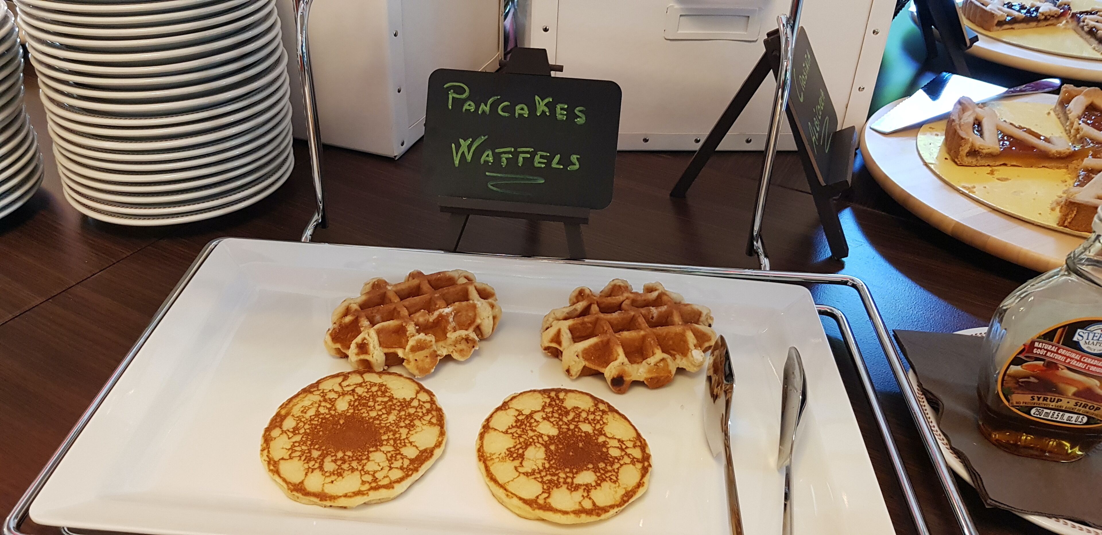 Ein Frühstückstisch mit Waffeln und Pfannkuchen auf einem weißen Teller, ein Schild mit der Aufschrift 'Pfannkuchen und Waffeln' sowie eine Sirupflasche.
