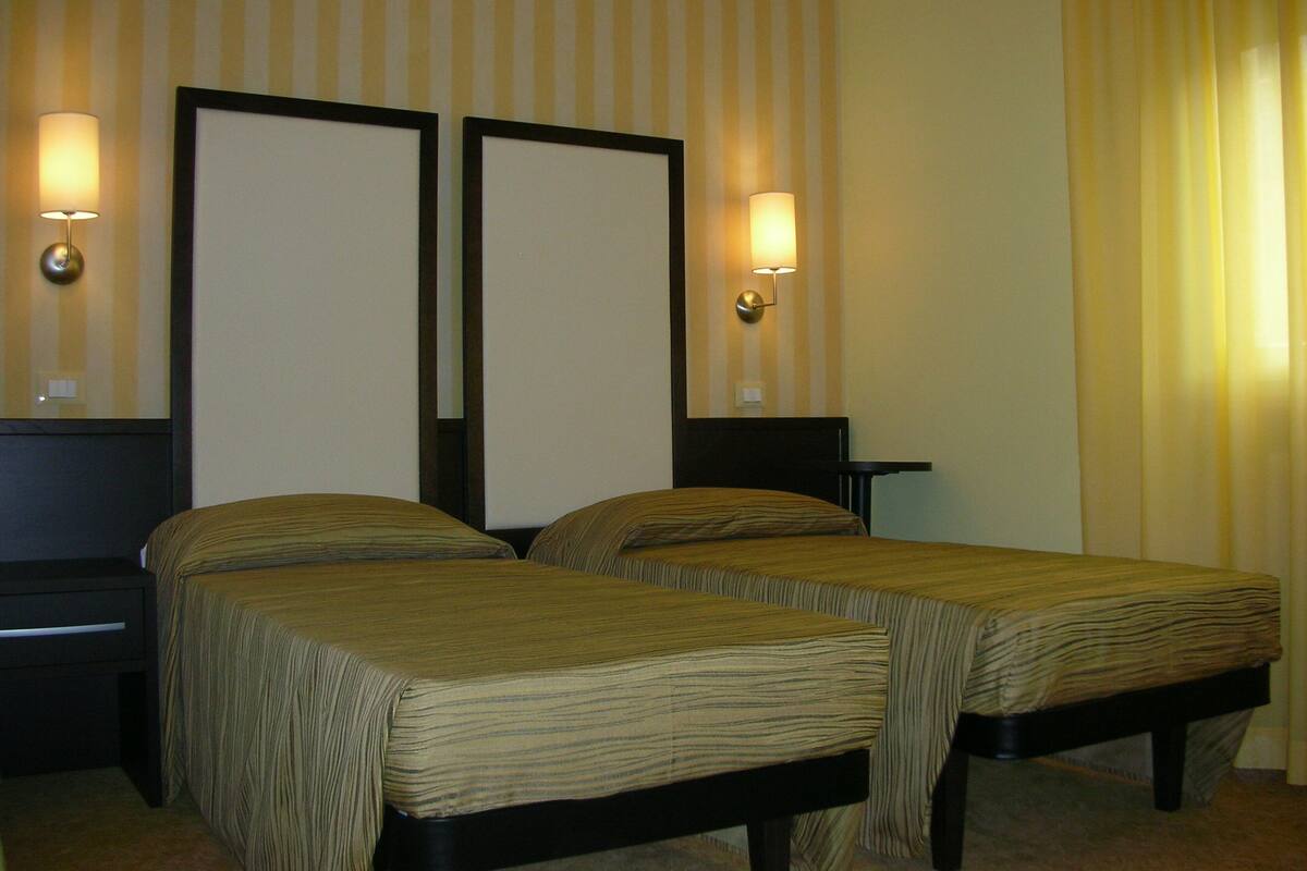 Quarto de hotel com duas camas de solteiro, mesa de cabeceira, luminária e iluminação embutida na parede.