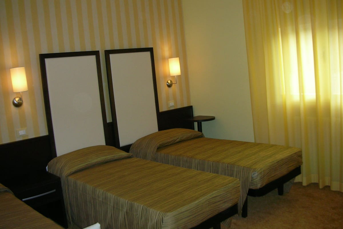 Quarto de hotel com duas camas de solteiro, uma mesinha e um abajur.