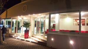 L’entrée d’un hôtel avec des portes en verre, un jardinier rouge et un espace détente avec des coussins rouges.