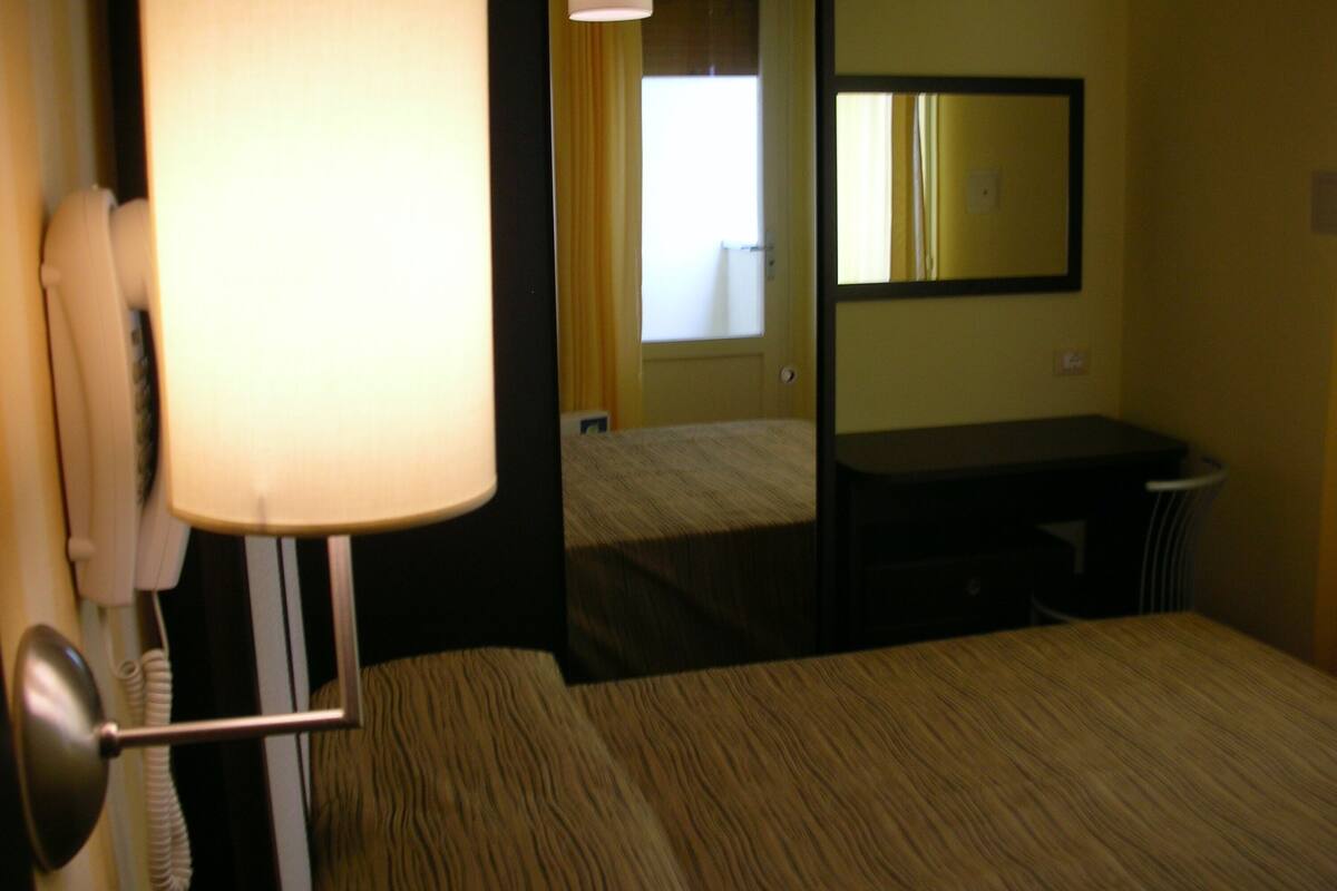 Quarto de hotel com cama, escrivaninha, abajur e espelho.