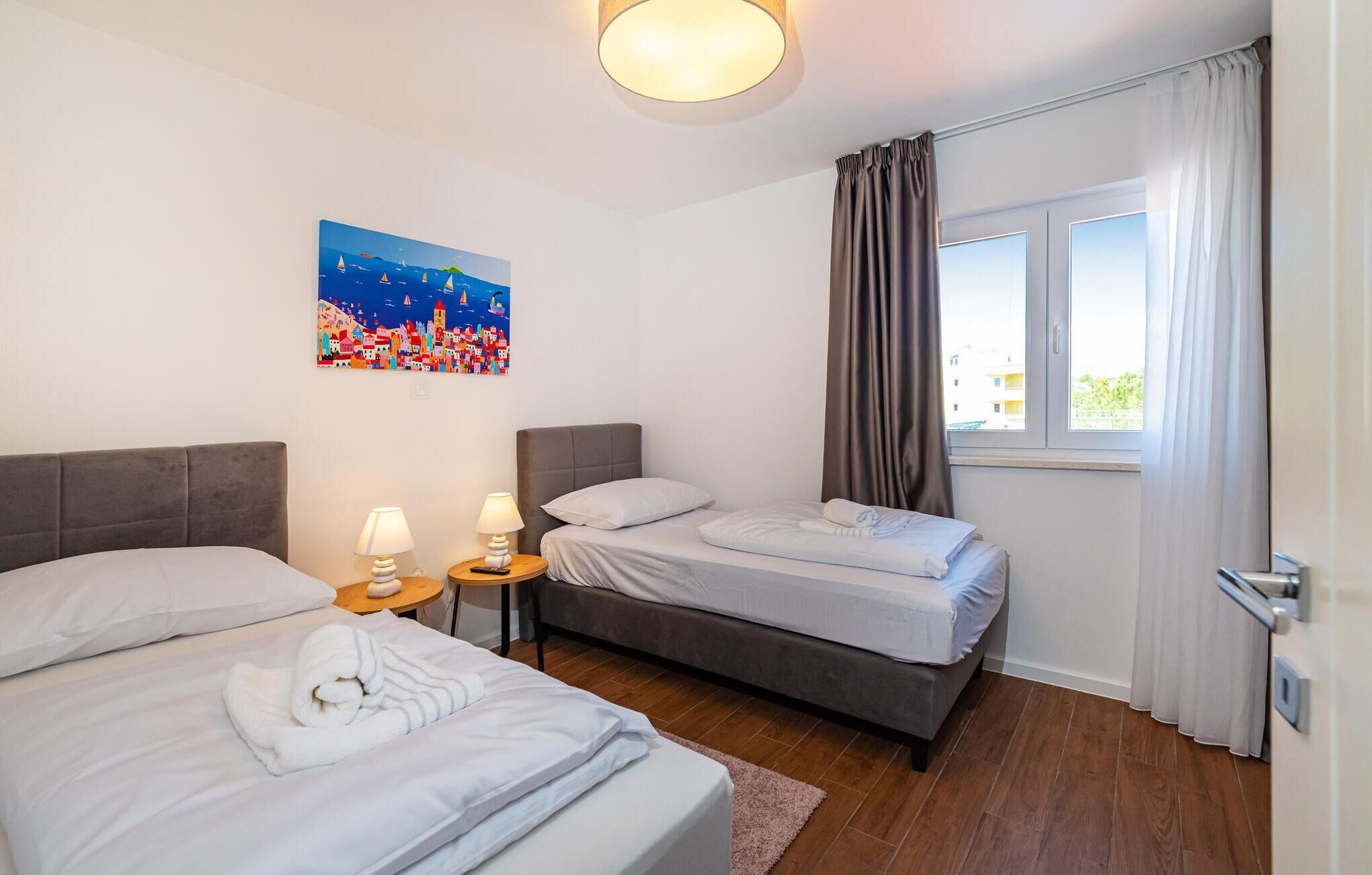 A hotel room with two beds, a painting of a coastal scene, a window with curtains, and a wooden floor.