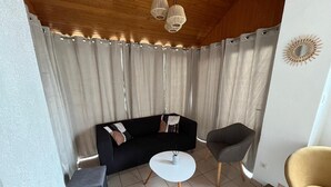 A modern living room with a black sofa, a grey armchair, a small white table, and a yellow chair. The room has wooden ceiling beams, a white wall, and a curtain-covered window.