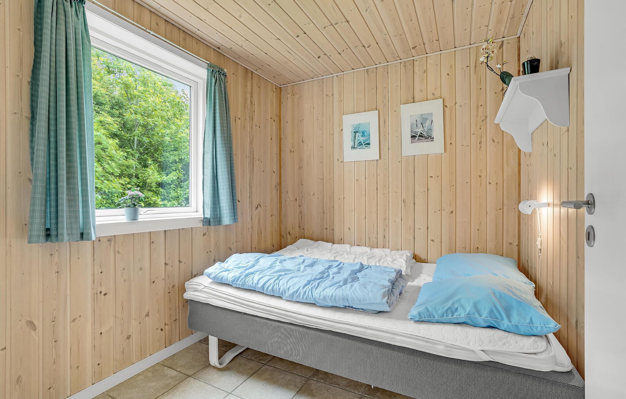 A room with wooden walls, a bed with blue bedding, a window with curtains, and two framed pictures on the wall.