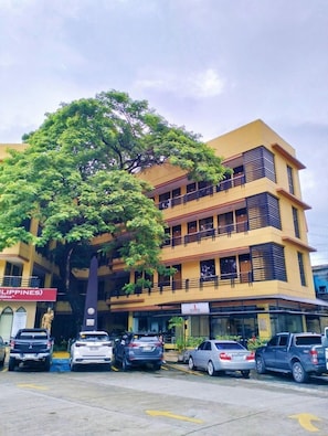 A multi-story building with a large tree in front, parked cars, and a statue. - Reddoorz Arzo Hotel And Resorts (Manila)