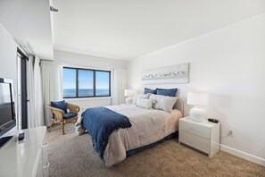 A hotel room with a large bed, a chair, a desk, a TV, a window with a view, and a wall-mounted artwork.