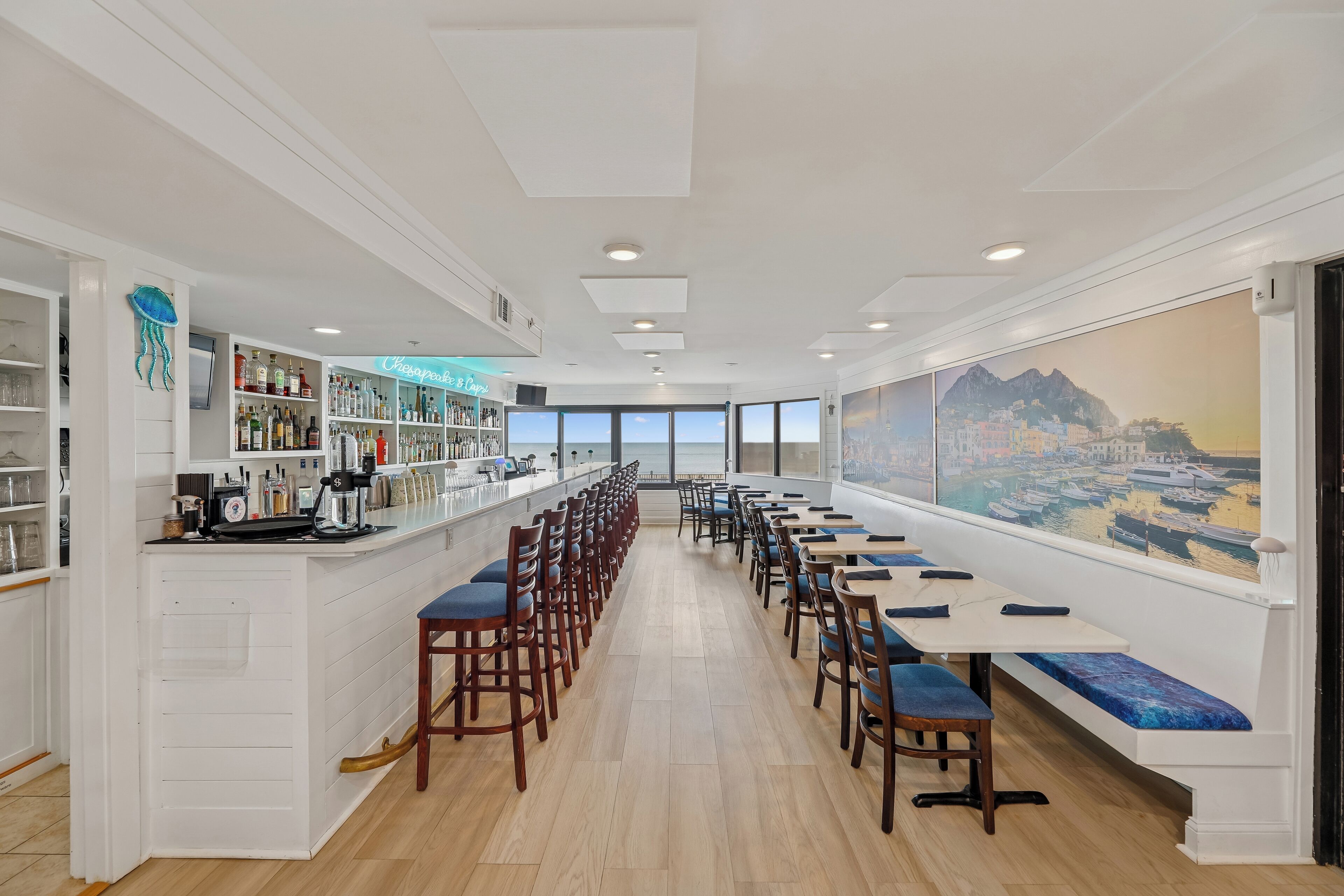 A long dining area with high tables and chairs, a bar on the left, and a large window with a cityscape view.