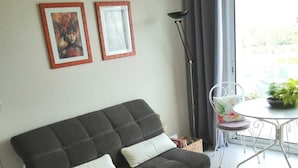 A living room with a dark grey sofa, two white cushions, a small round table, and a patterned rug. There are two framed pictures on the wall, a floor lamp, and a window with curtains. - La Rochelle T2 35m2 Avec Terrasse Dans Résidence de Standing (La Rochelle)