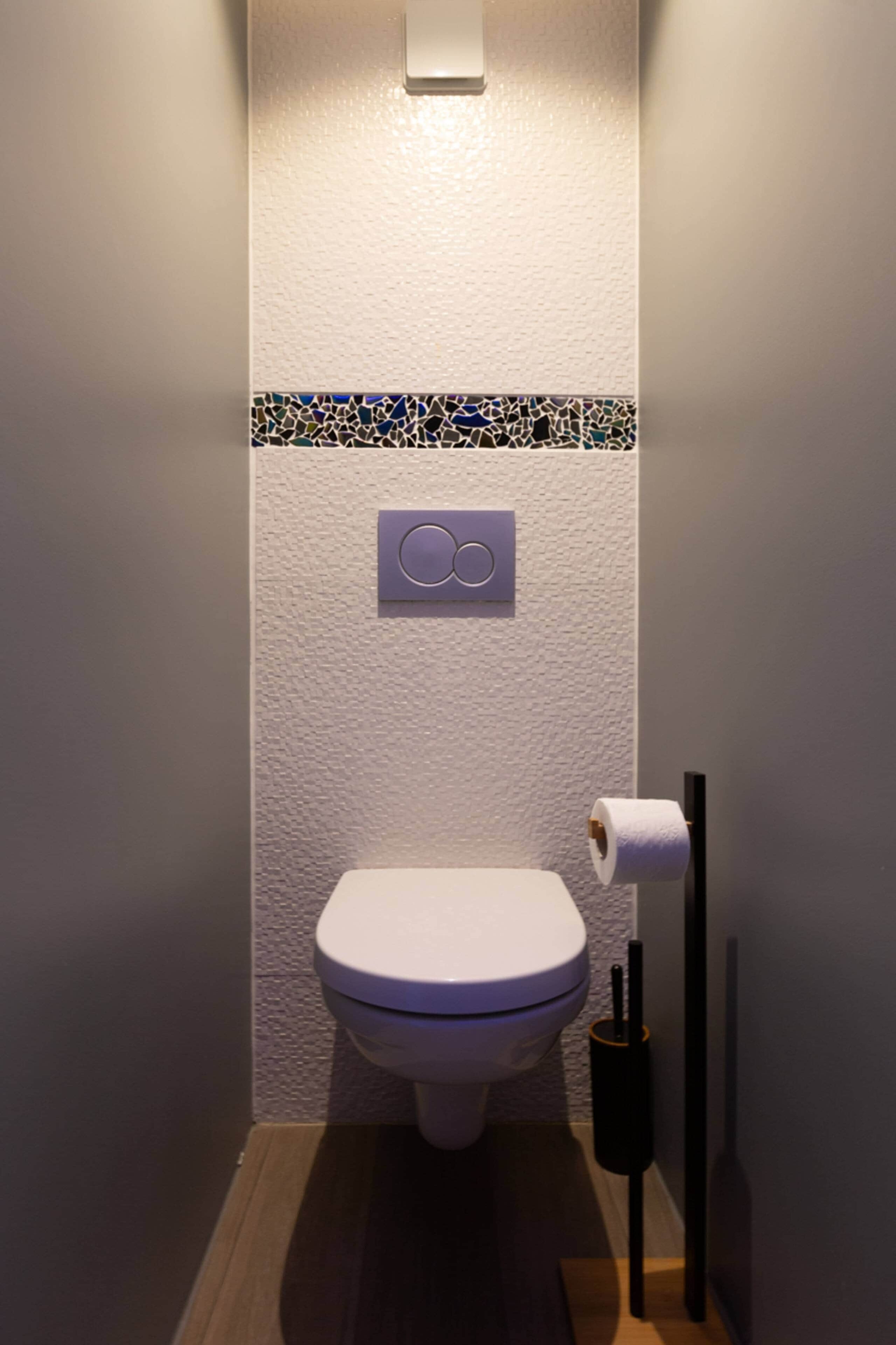 A small, modern bathroom with a white toilet, a toilet paper holder, and a wall-mounted flush button.