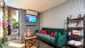 Un soggiorno moderno con un divano verde, un tavolino da caffè e una TV montata a parete. Si vede una libreria con un opuscolo di benvenuto e uno scaffale con asciugamani.