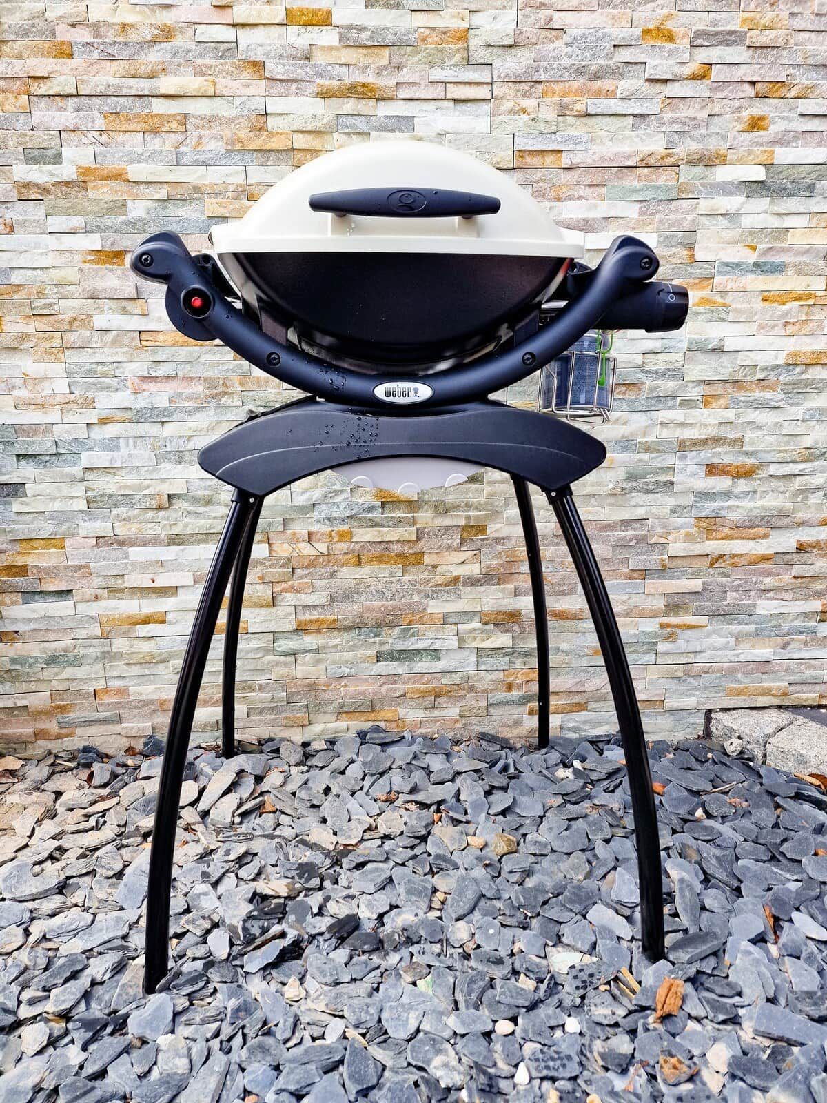 A portable barbecue grill with a black handle and a logo, standing on a black metal stand, against a stone wall background.