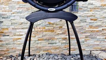 A portable barbecue grill with a black handle and a logo, standing on a black metal stand, against a stone wall background.