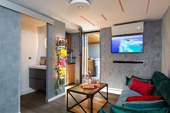A modern living room with a flat-screen TV, a sofa with colorful cushions, and a table with a bottle of wine.