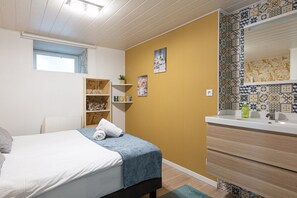 Un dormitorio con cama, mesita de noche, estante con objetos decorativos, ventana y baño con lavamanos y espejo.